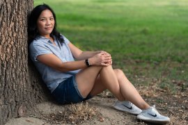 A photo of an Asian American woman seated outside, leaning against a tree.