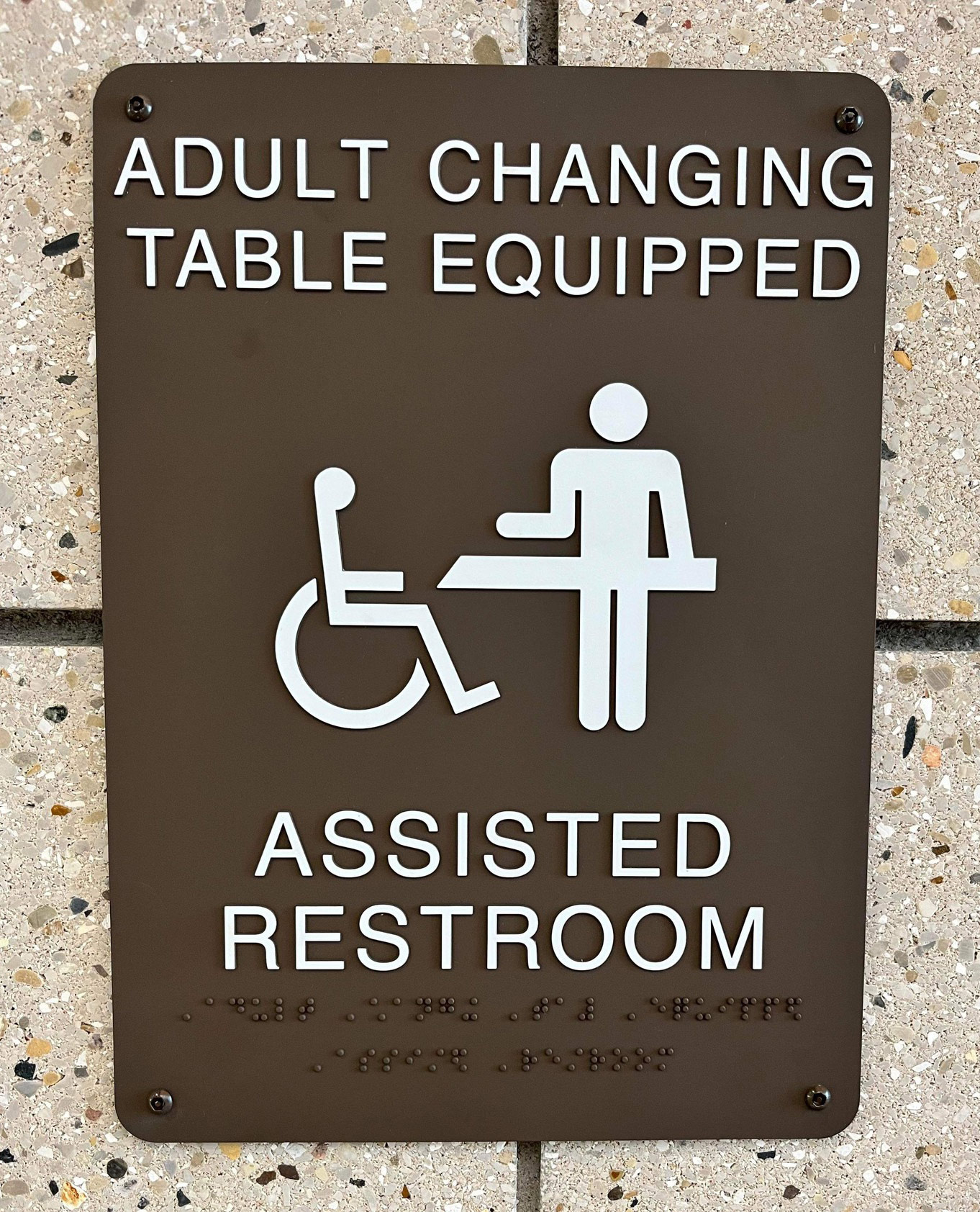 A photo of a sign outside a bathroom that shows a person in a wheelchair and a person preparing to use an adult-size changing table.