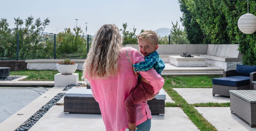 A woman in a pink shirt walks away from the camera while holding a young boy on her right him. The boy looks behind her at the camera.