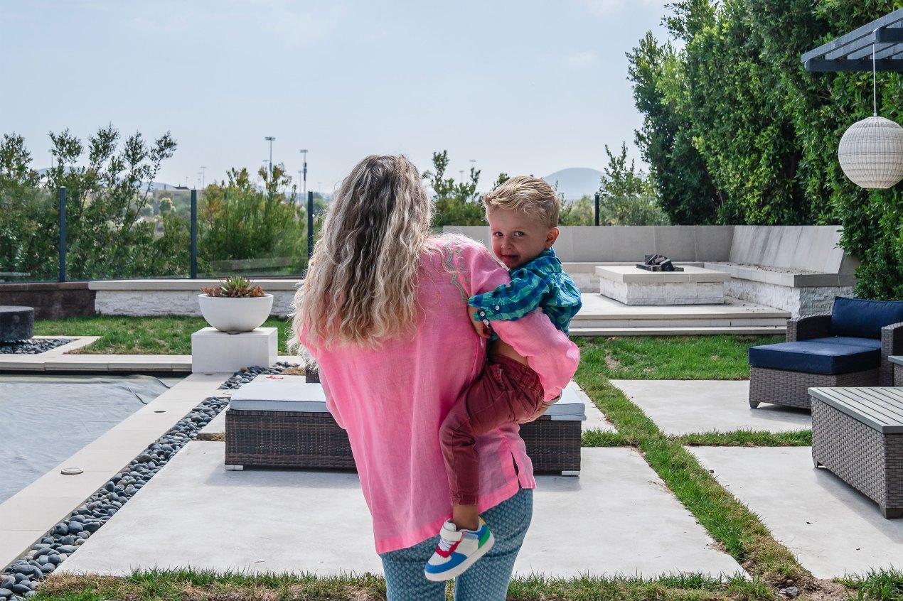 A woman in a pink shirt walks away from the camera while holding a young boy on her right him. The boy looks behind her at the camera.