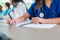 An unrecognizable medical student sits at a table in a lecture hall and writes in her class workbook.