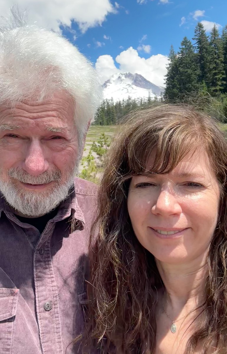 A man with white hair and a beard stands next to a brown-haired woman