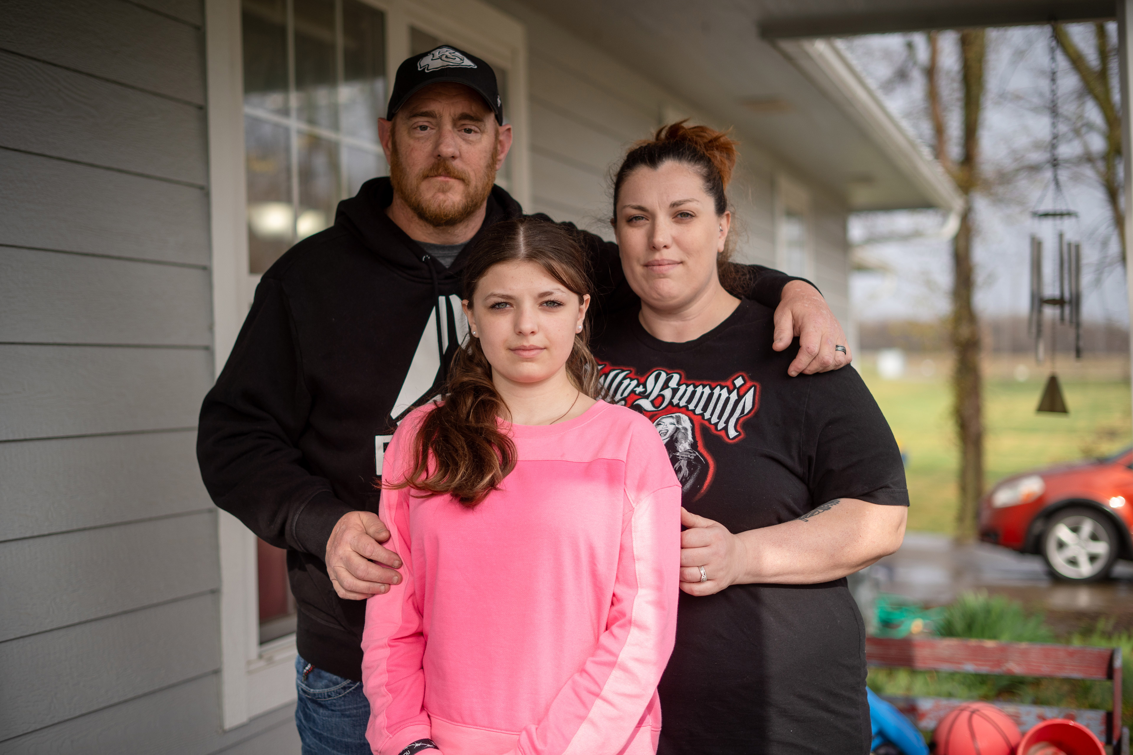 A man, a woman, and a young girl pose for a photo as they stand on the porch in front of a house