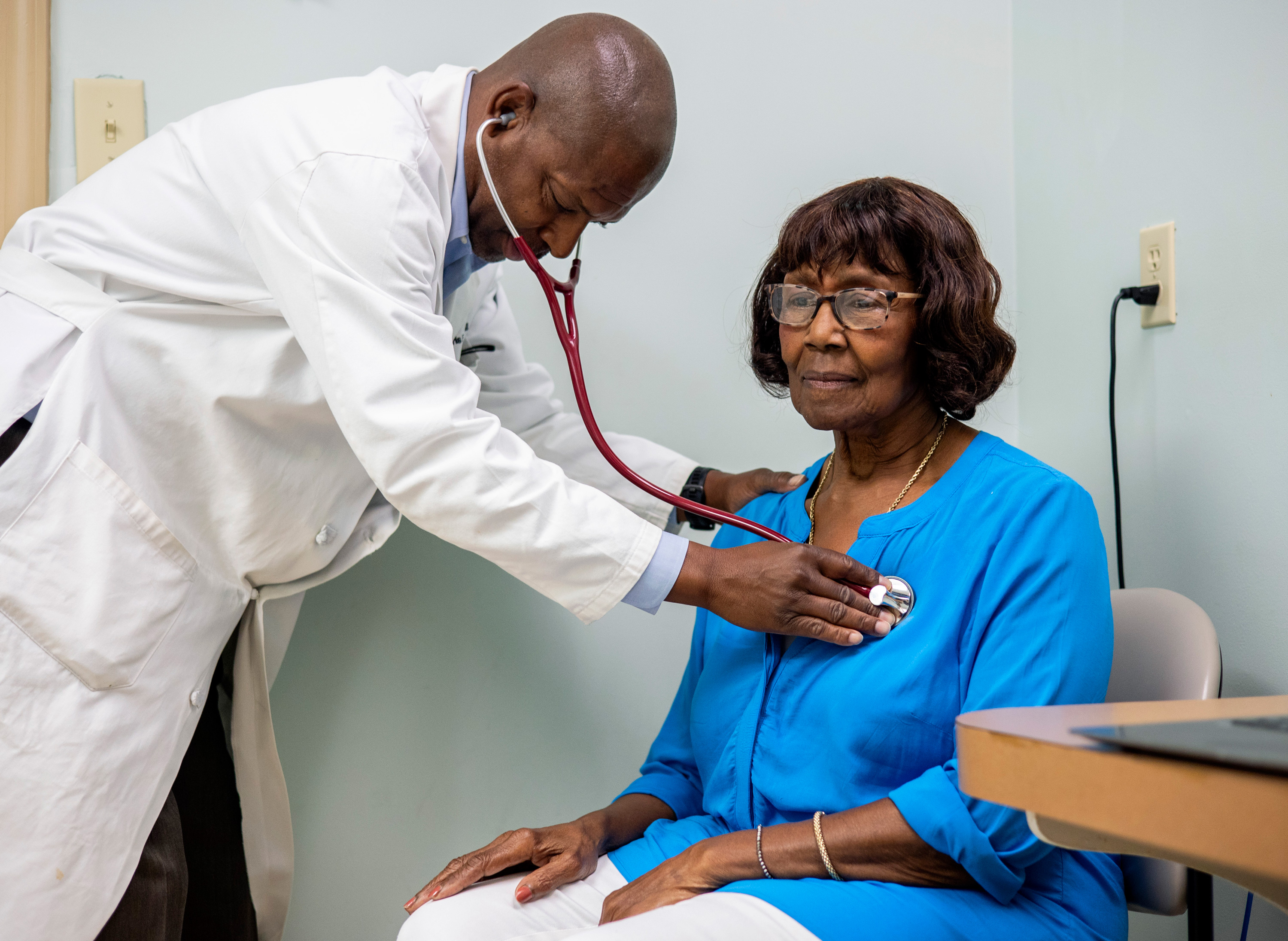 A photo of a Black doctor using his stethoscope on his patient, an older Black woman.