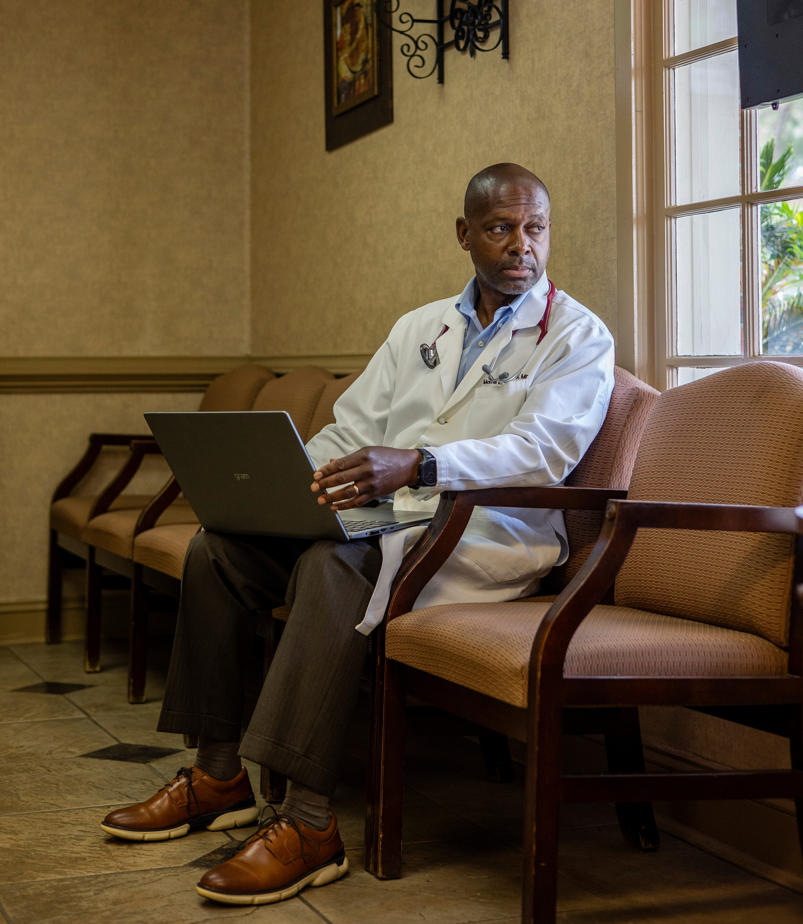 A photo of a Black doctor sitting in his office, looking to the right toward a window.