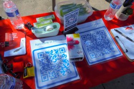 A photograph of an outdoor table with Naloxone supplies including a box of Narcan, information clips, bottled water, information pamphlets, and QR codes of where to find Naloxone in Denver, CO, and Colorado at large.