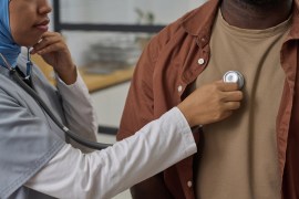 A female primary care doctor examines a male patient using a stethoscope.