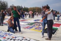 Groups of people view large quilt panels on the ground. In the foreground, two men hug, and nearby, a man puts his arm around the shoulders of another kneeling beside a panel.