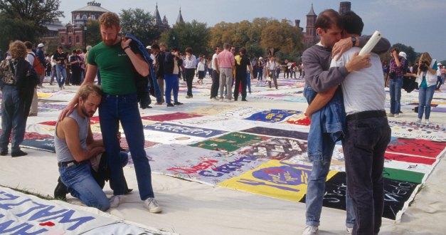 Groups of people view large quilt panels on the ground. In the foreground, two men hug, and nearby, a man puts his arm around the shoulders of another kneeling beside a panel.