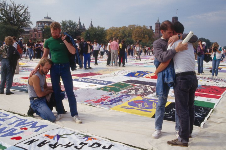 Groups of people view large quilt panels on the ground. In the foreground, two men hug, and nearby, a man puts his arm around the shoulders of another kneeling beside a panel.