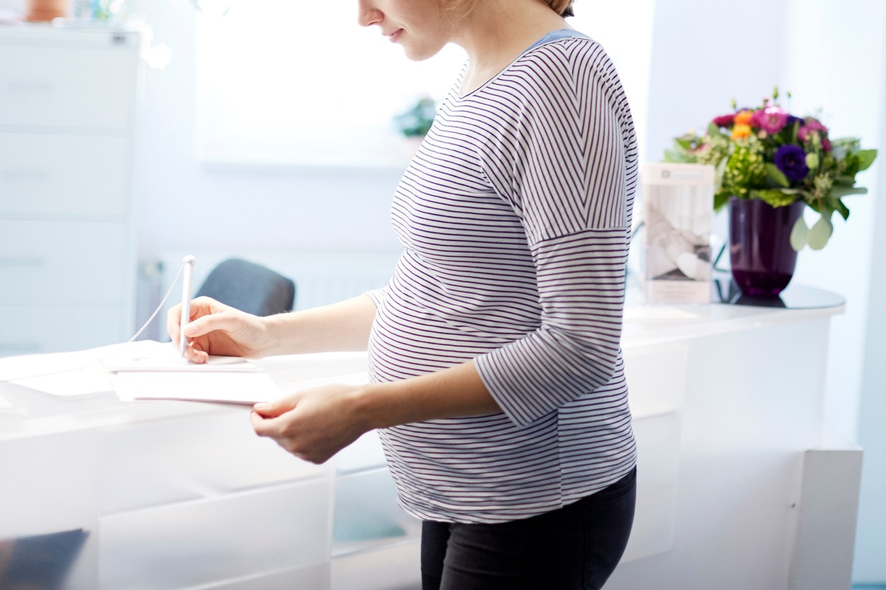 A pregnant woman is at reception for doctor's office, signing a piece of paper.