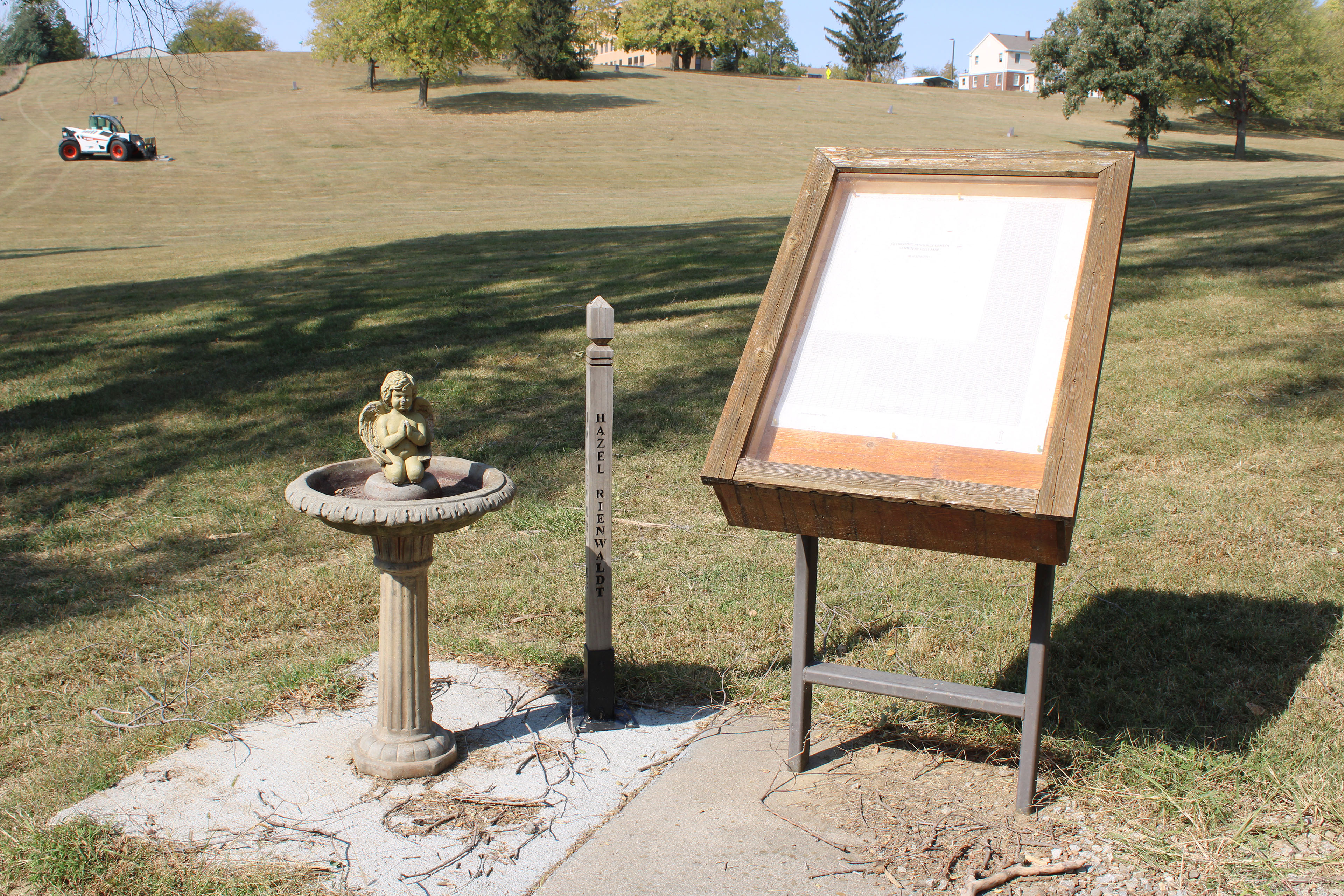 In a sunny cemetery, a large wooden sign is posted beside a birdbath with a stone angel in its center.