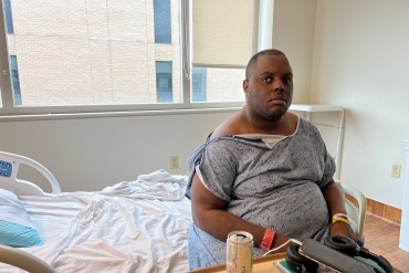 A man wearing a hospital gown is sitting on a bed in a hospital room alone.