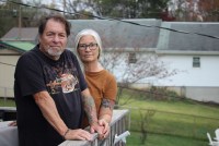 A portrait of a husband and wife standing on their porch. They face the camera with a serious expression.