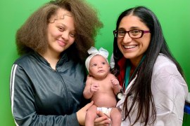 Two women smile at the camera while holding a baby with a white bow on her head