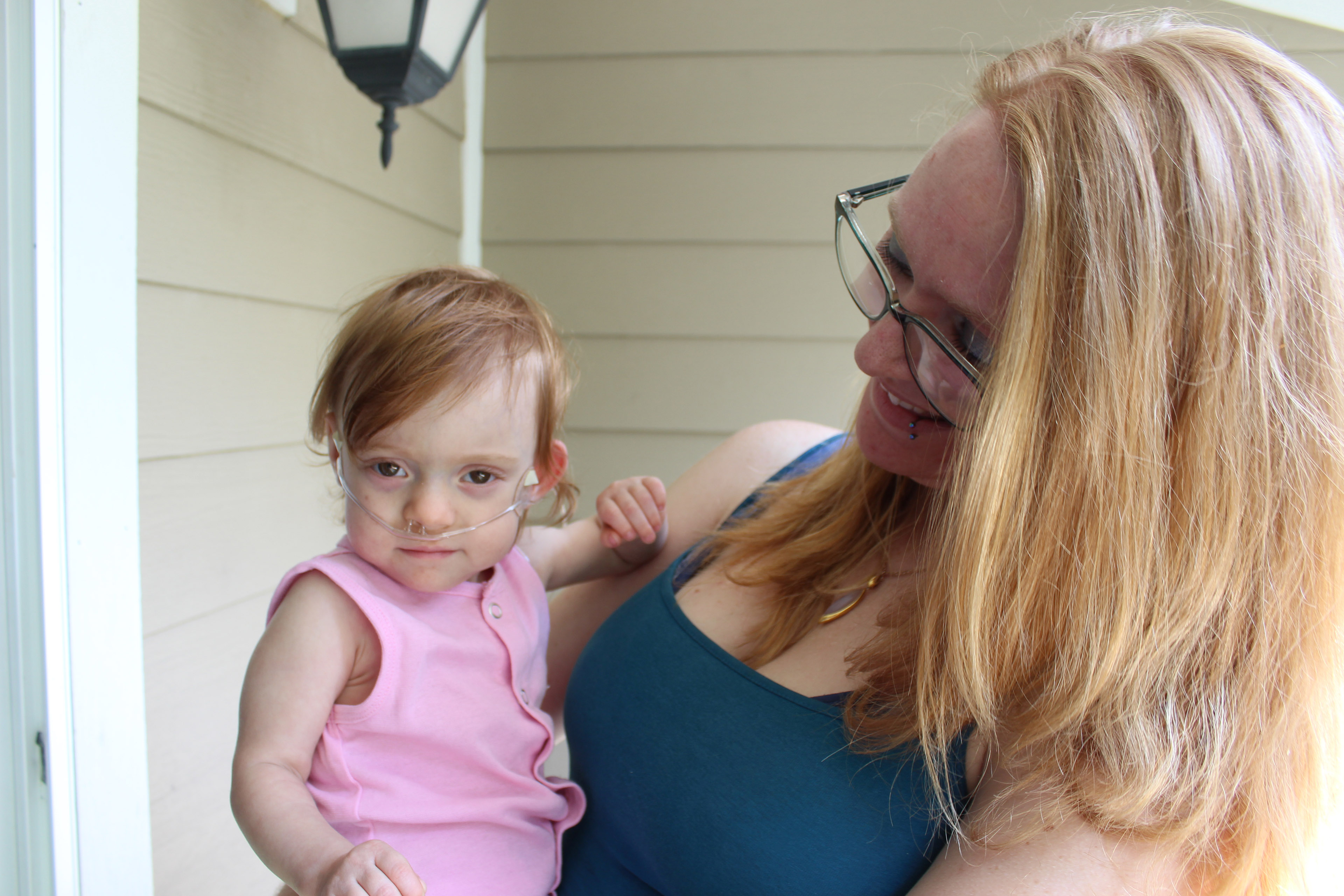 A woman wearing glasses and a blue tank top smiles at the baby she's holding, who has red hair and is wearing a pink outfit