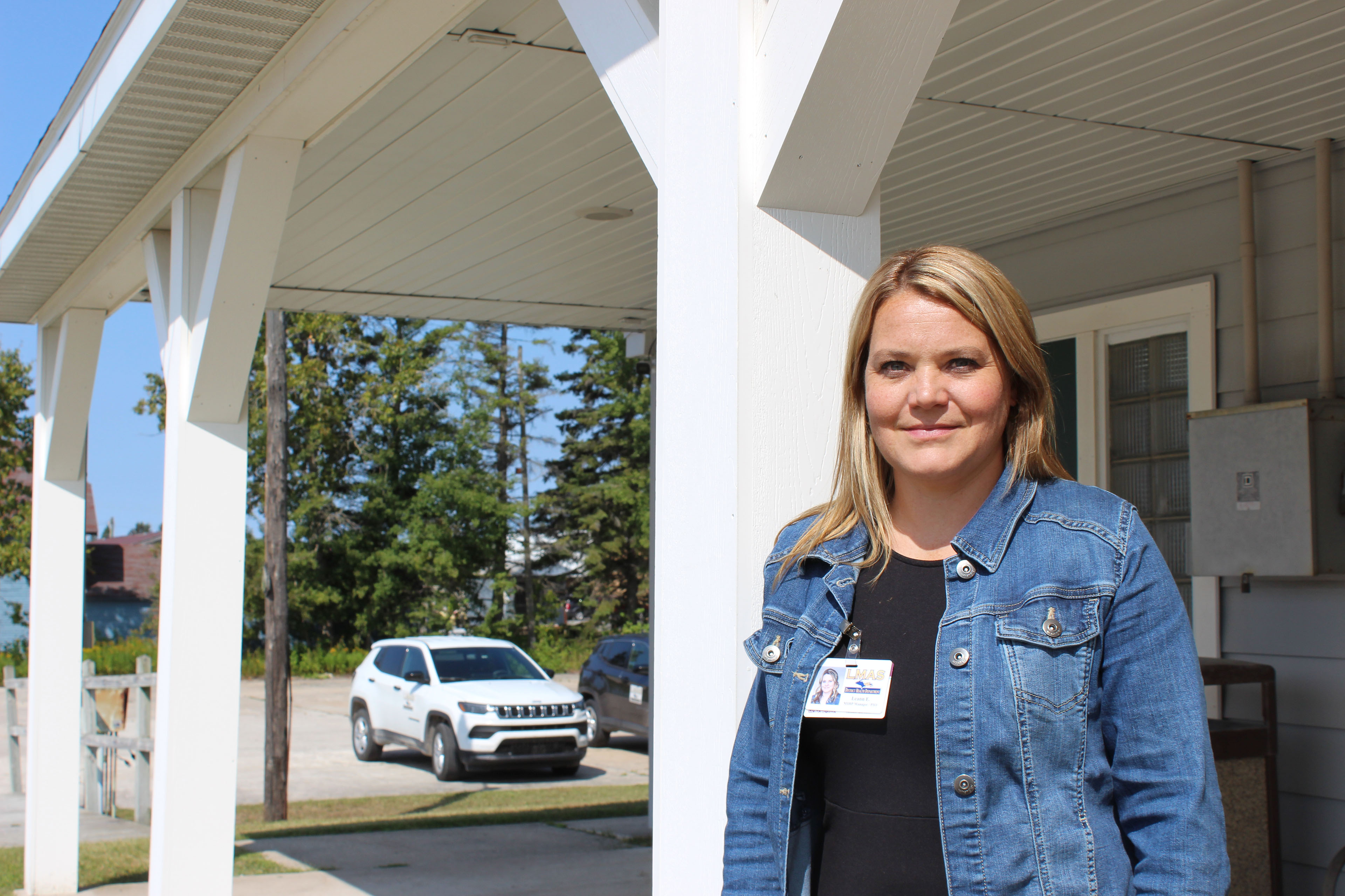 A woman wearing a black shirt and denim jacket leans against a white post and smiles at the camera