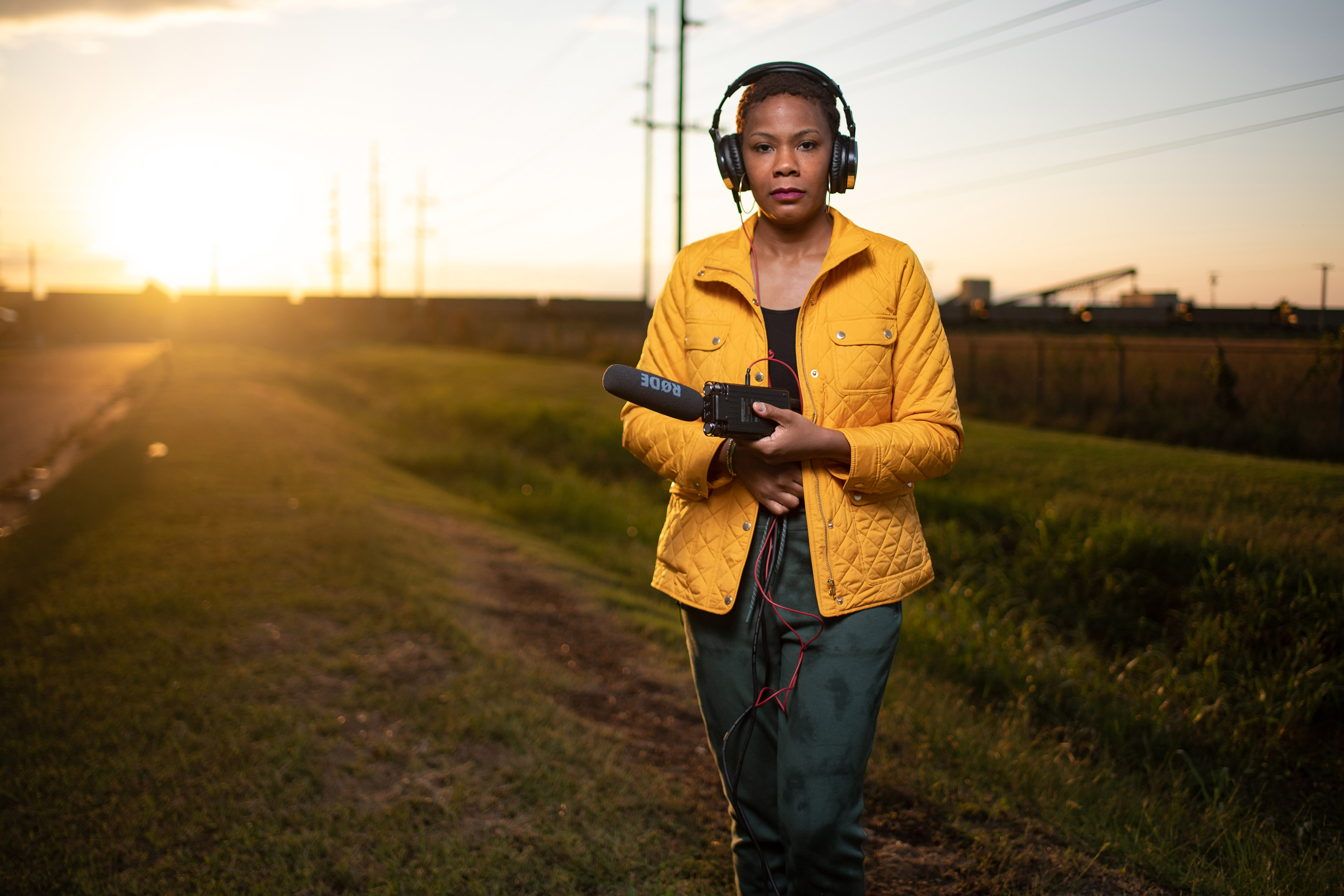 Cara Anthony stands wearing and holding an audio kit in a field at sunset.