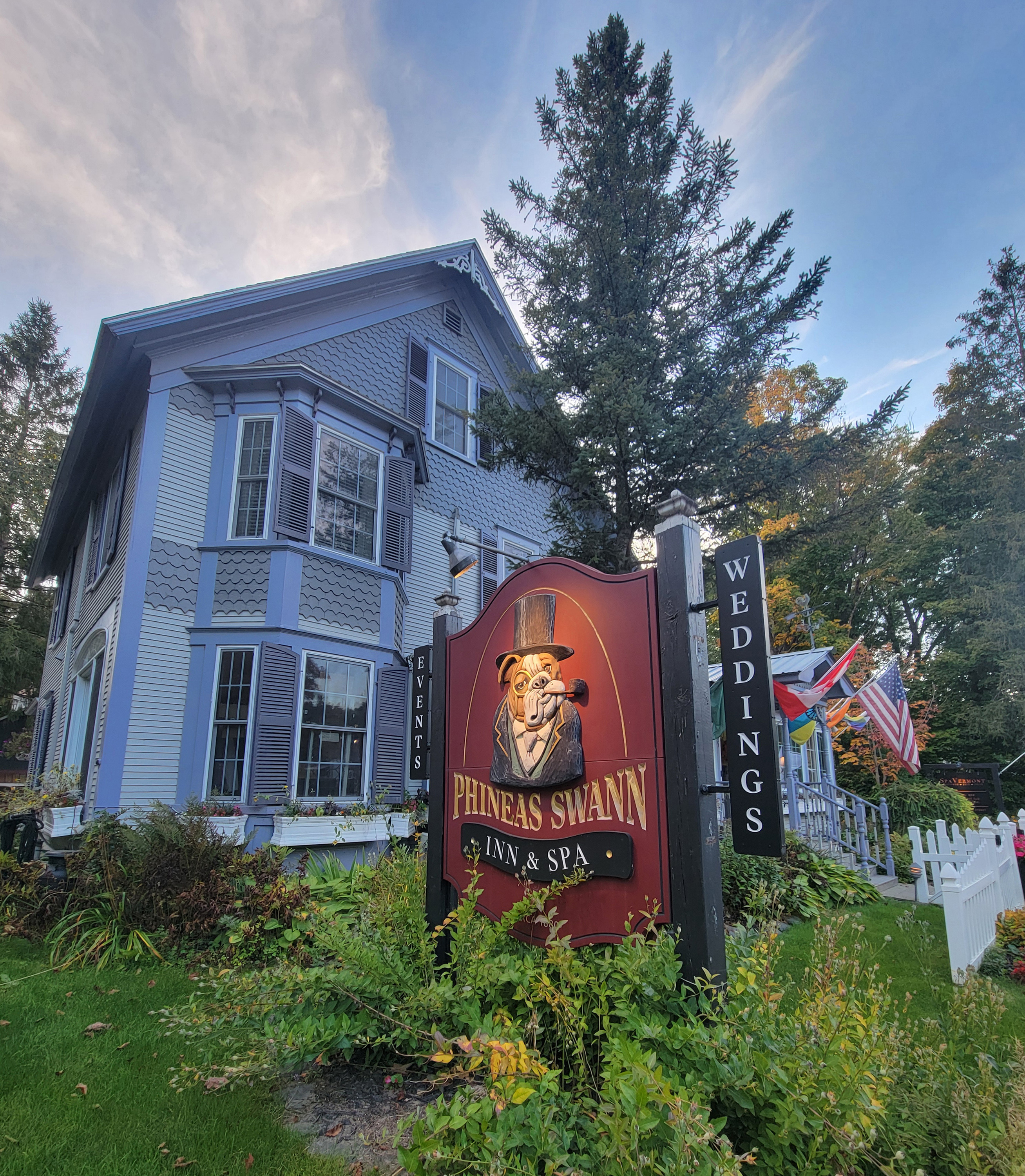 A photo of the outside of an inn. A sign outside it has its name above an image of a dog wearing a top hat and monocle, smoking from a pipe.