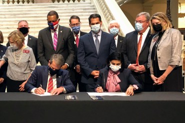 Georgia Gov. Brian Kemp (left) and Centers for Medicare & Medicaid Services Administrator Seema Verma (right) sit at a table facing the camera while they sign papers. Nine other individuals in business attire stand in a row behind them. Everyone is wearing face masks.