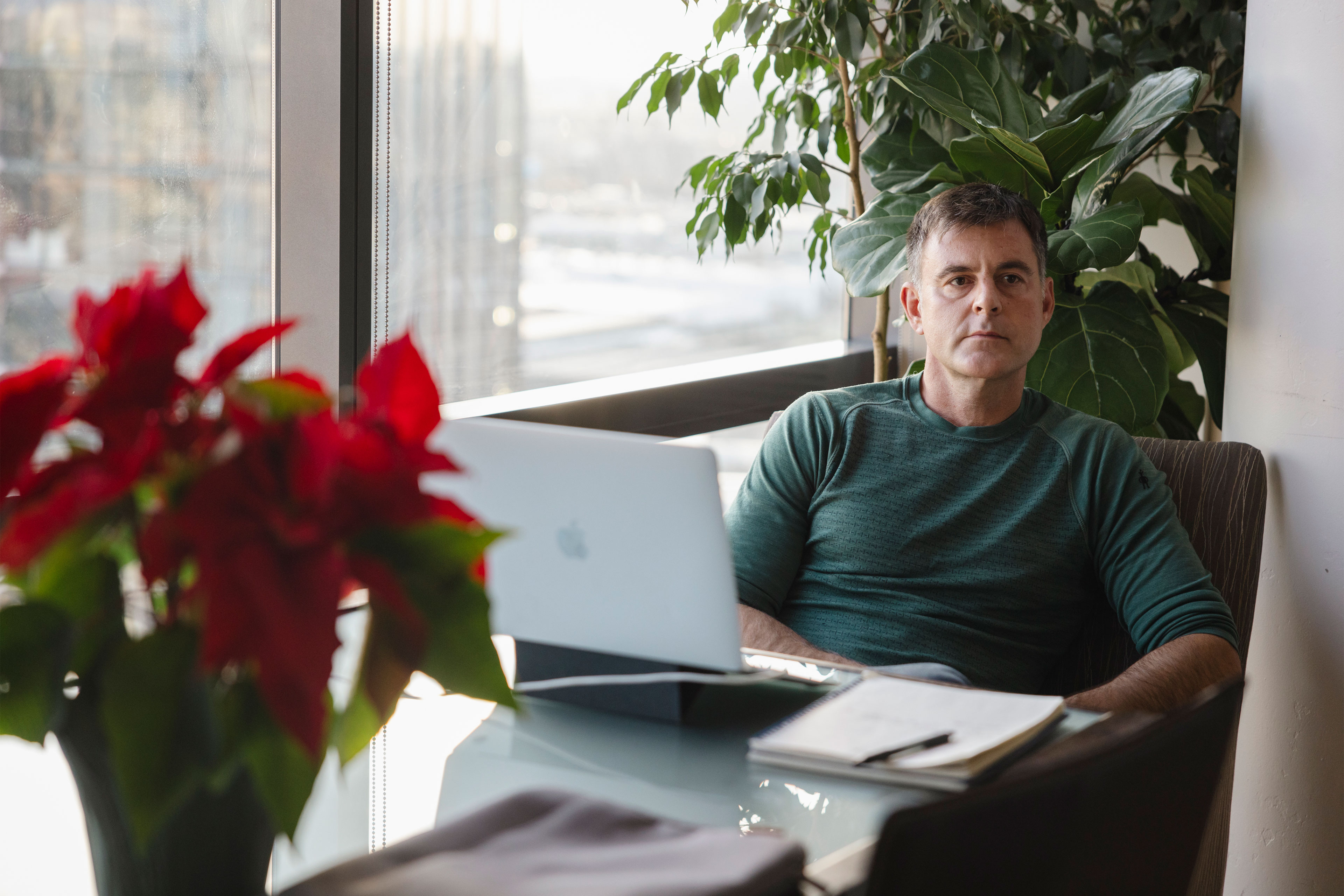 A man sits at a table near a window. A poinsettia plant, laptop, and notebook with pen rest on the table in front of him.