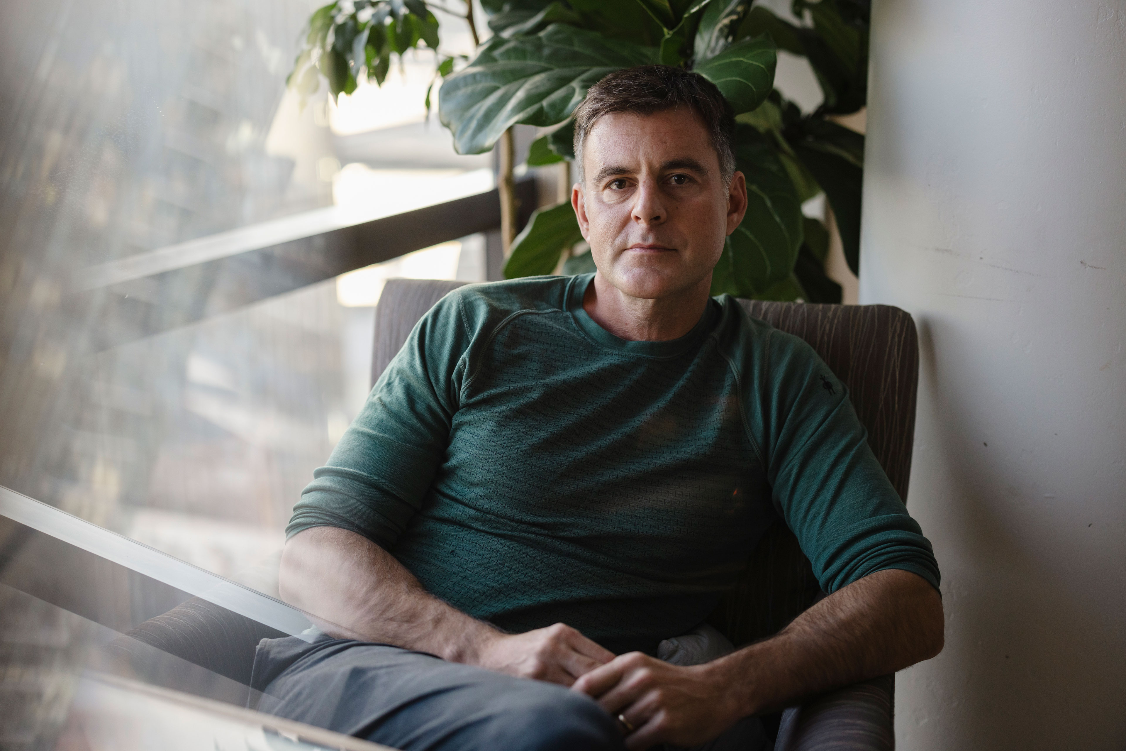 A man sits in an armchair near a window and looks at the camera.