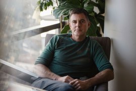 A man sits in an armchair near a window and looks at the camera.