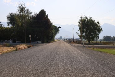 A landscape photograph of a dirt road in a rural setting. The road extends into the distance.