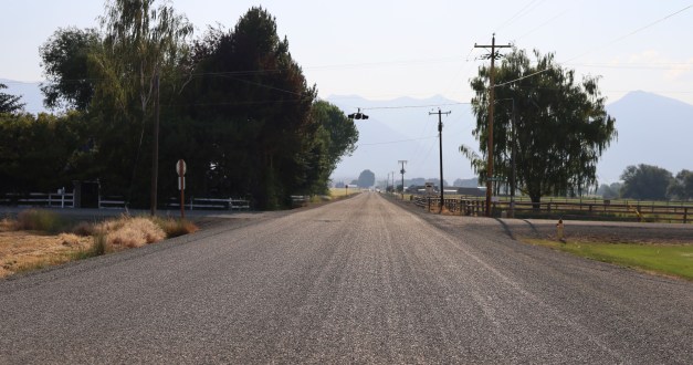 A landscape photograph of a dirt road in a rural setting. The road extends into the distance.
