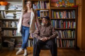 Two people are photographed in front of bookshelves. A seated man with a bushy gray beard looks off-camera to his right while a woman stands, leaning against the chair, and looks at the camera.