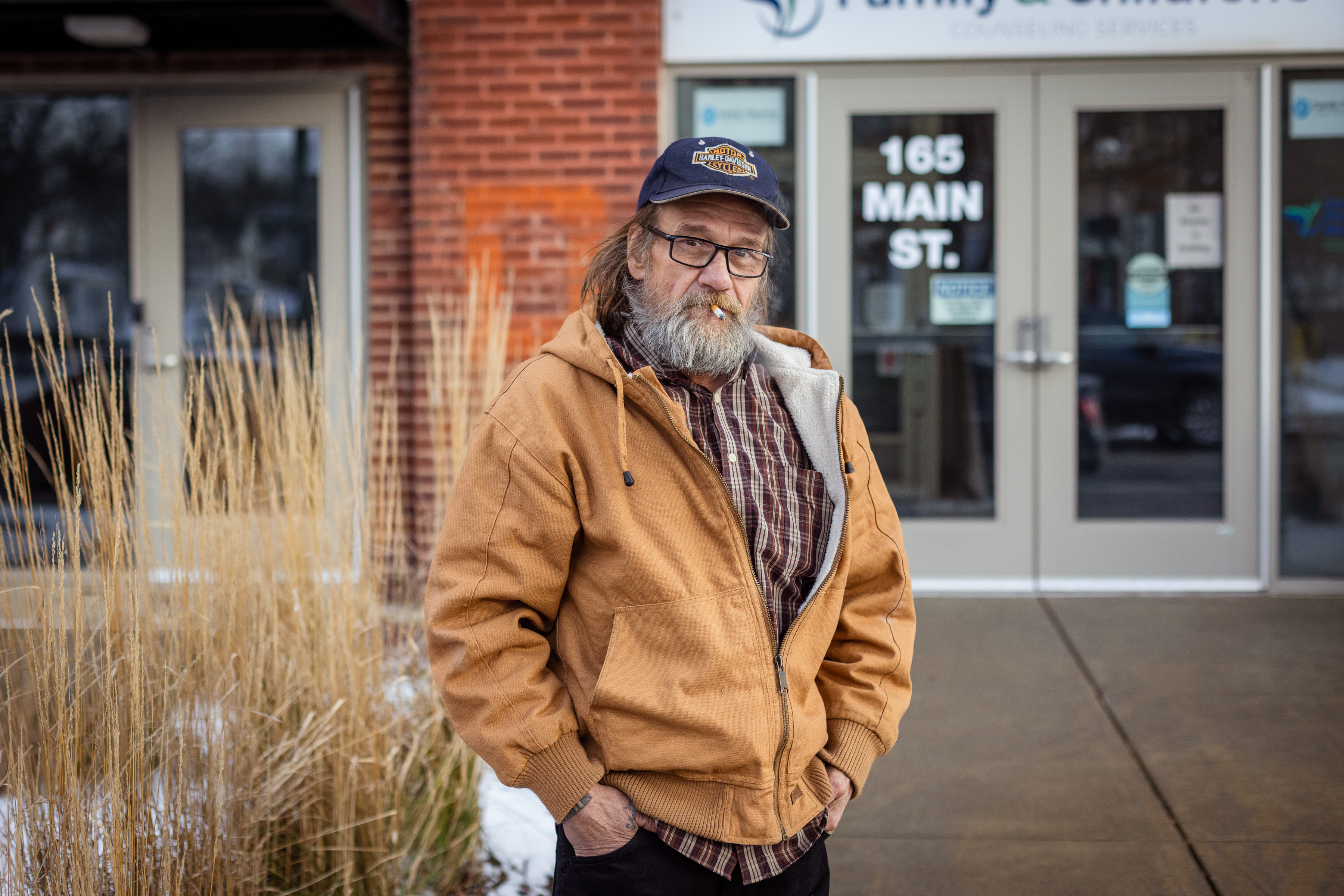 A man with a bushy gray beard in a jacked and cap stands outside a building and smokes a cigarette while looking at the camera.