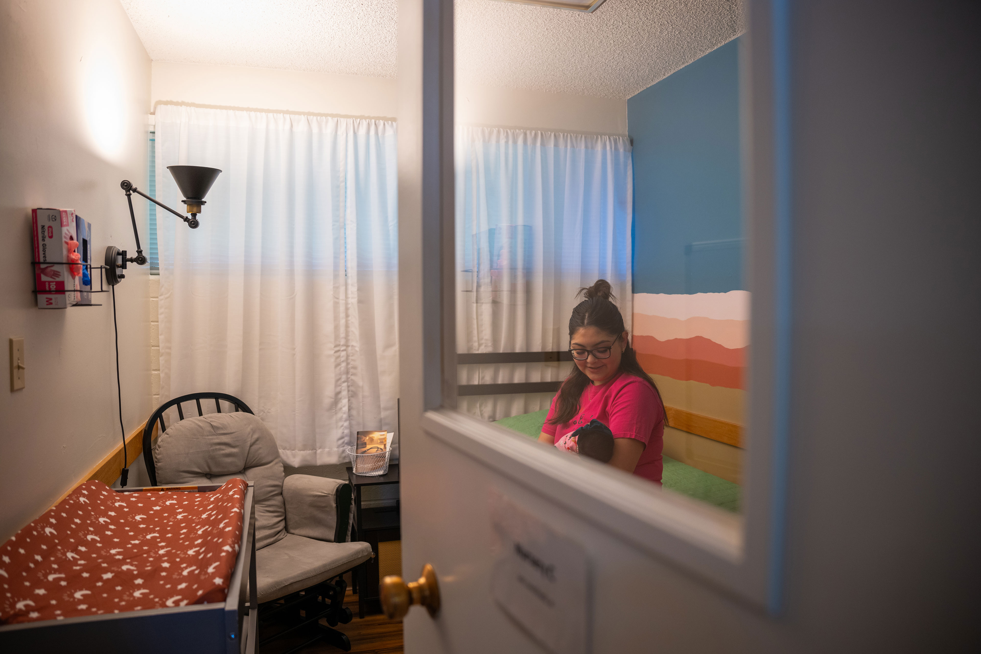 A view of a room through the window in the door with a bed, armchair and changing table. A woman sits on the bed and looks down at the infant in her arms.