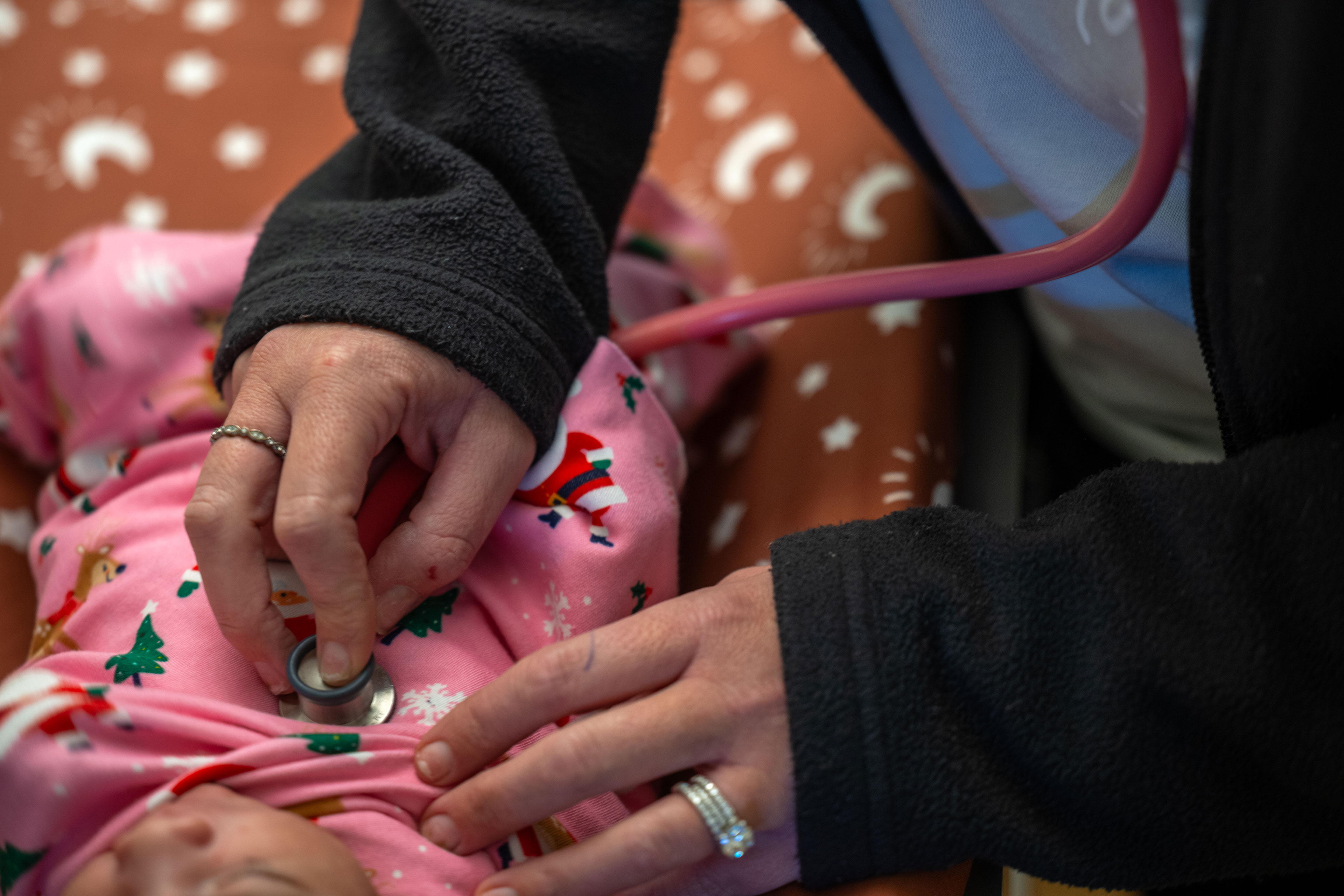 A close-up image of a nurse holding a stethoscope to baby's chest. The unidentifiable baby is wrapped in a pink cloth with a pattern of  Santa, reindeer, trees, and snowflakes.