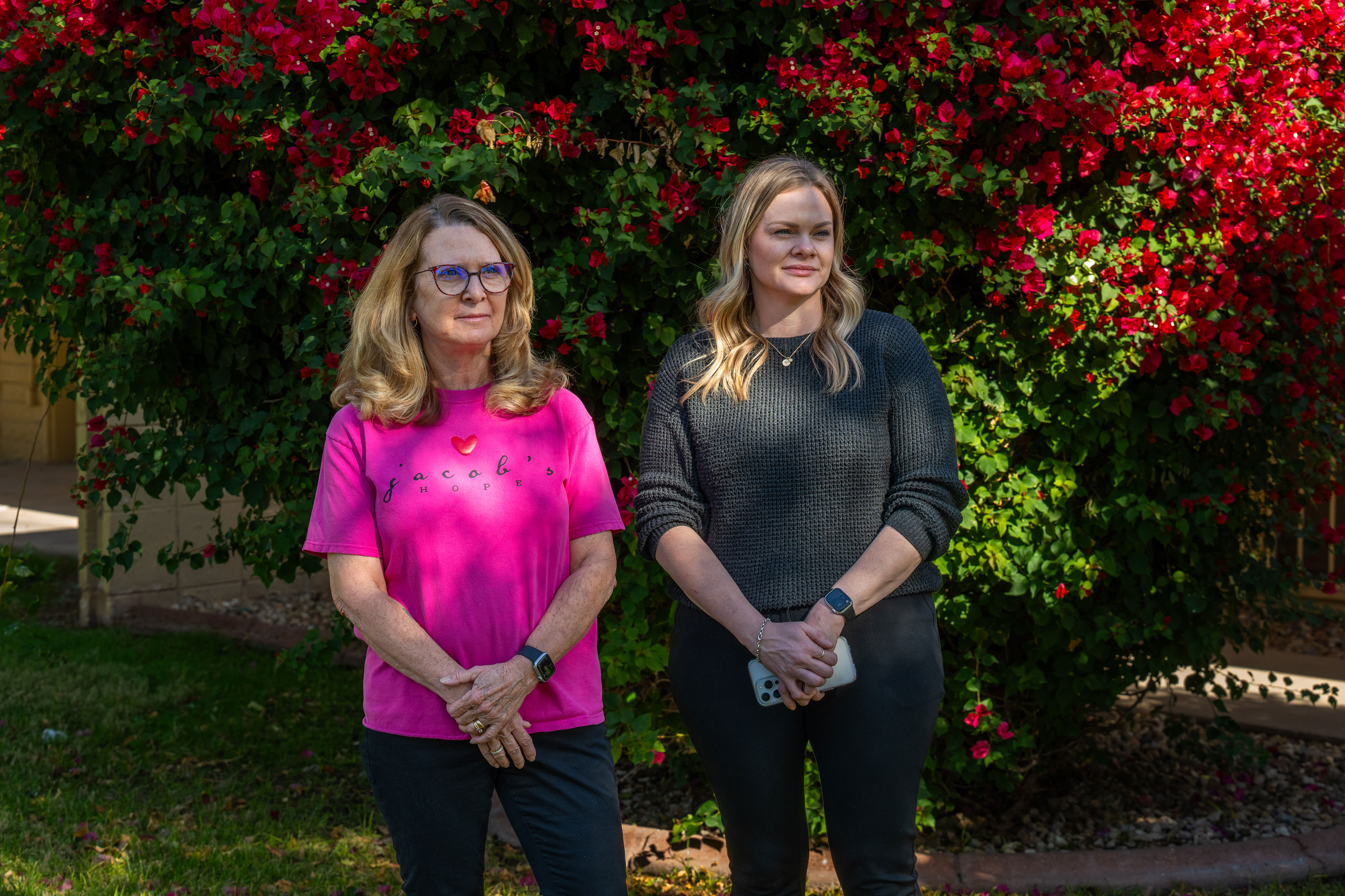 Two women stand in front of a large flowering bush with their hands clasped. One wears a pink shirt that reads "Jacob's Hope" and the other wears a dark gray sweater.