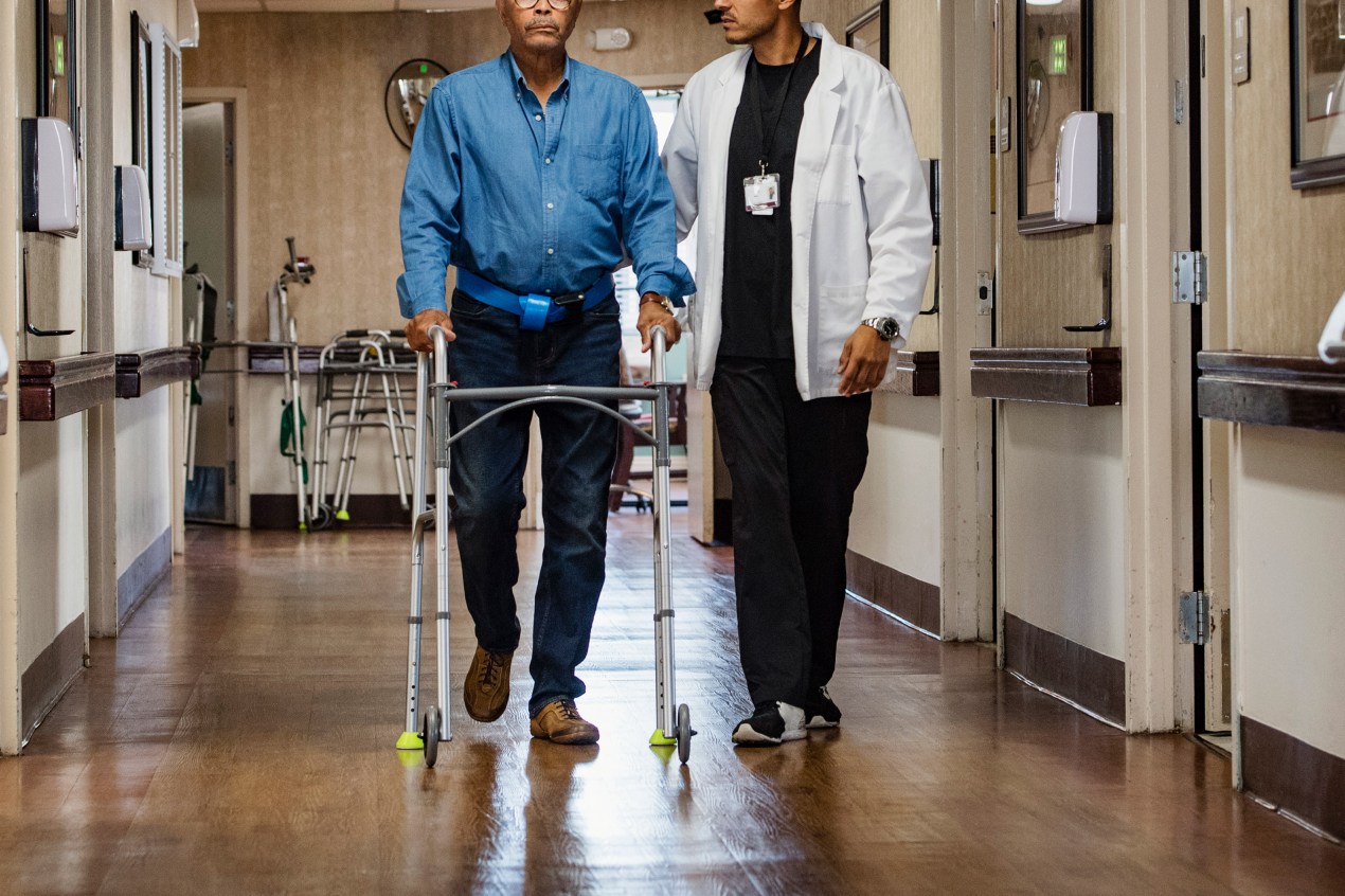 A photo of a nursing home patient being helped walking down a corridor by a staff member.