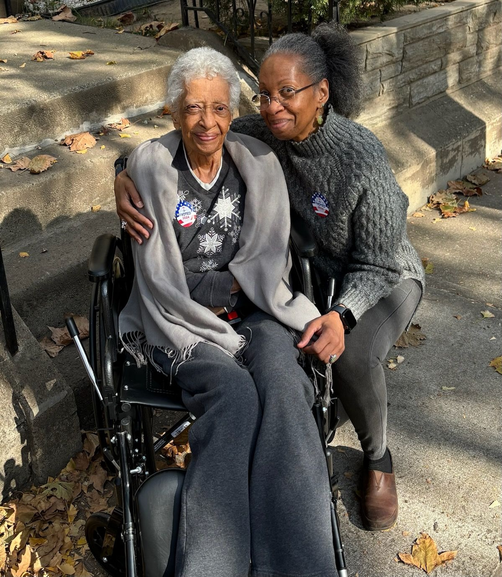 A portrait of two women side-by-side. On the viewer's left is a senior woman in a wheelchair. On the right is a middle-aged woman who has her arm around the senior woman as she kneels beside her. They are both smiling warmly.