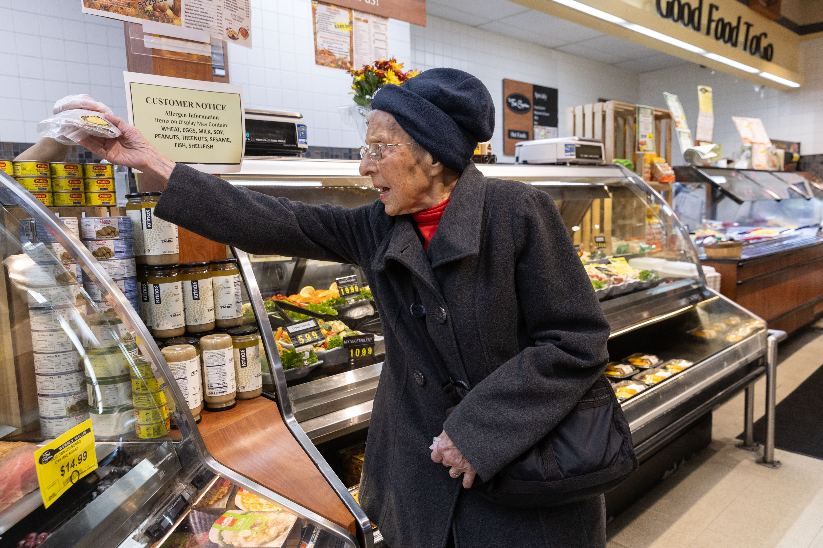 A photo of a senior woman wearing winter clothes picking up food from a deli counter.