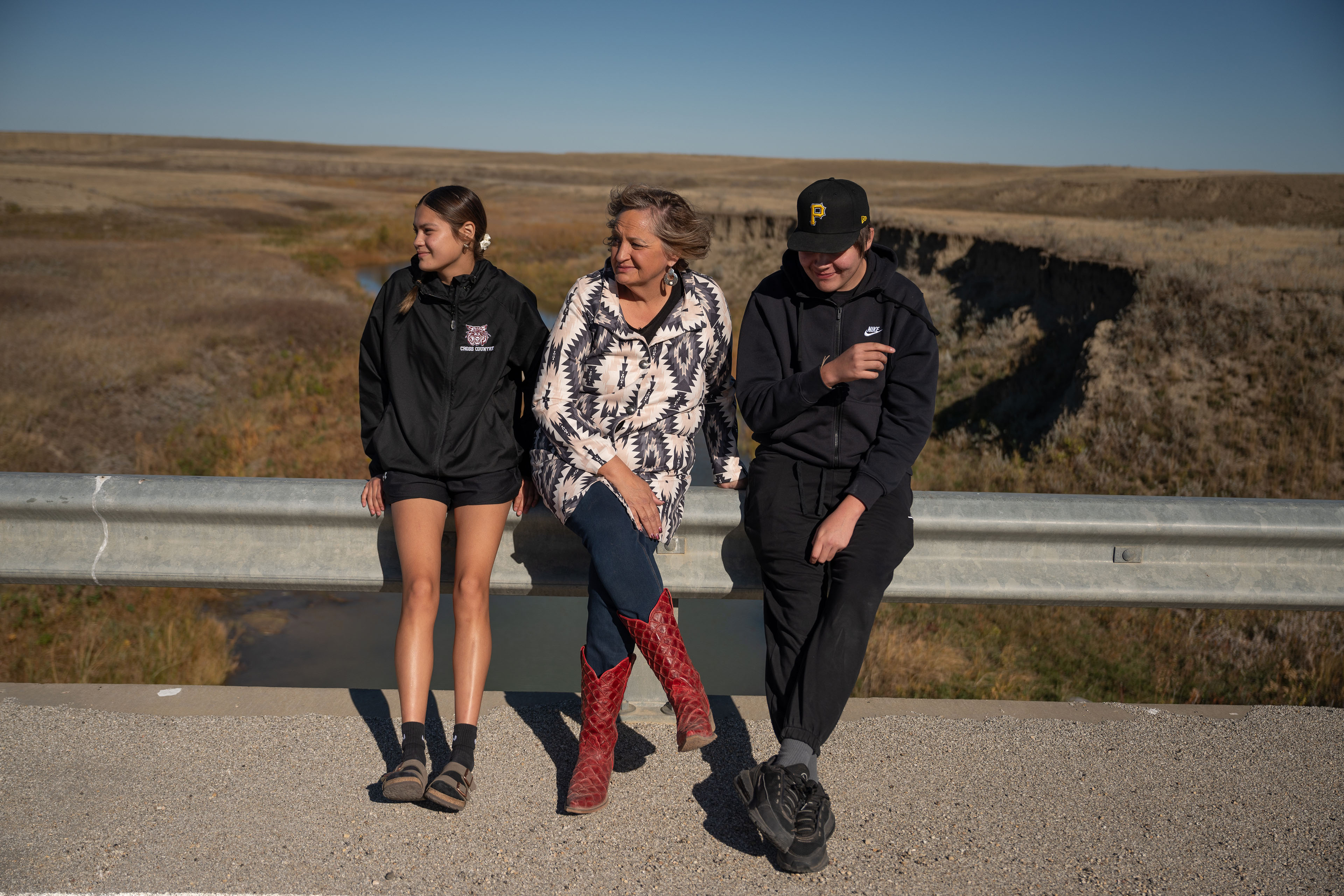 A photograph of a mother with her two children. Her daughter is on the viewer's left and her son is on the viewer's right, while she is in the center. The family is outside near a grassy field on a sunny day.