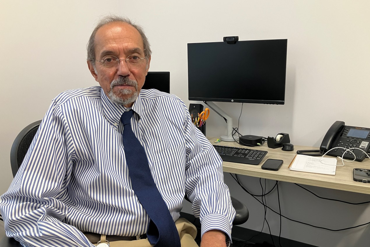 An older man in a navy striped shirt and a navy tie sits at a desk