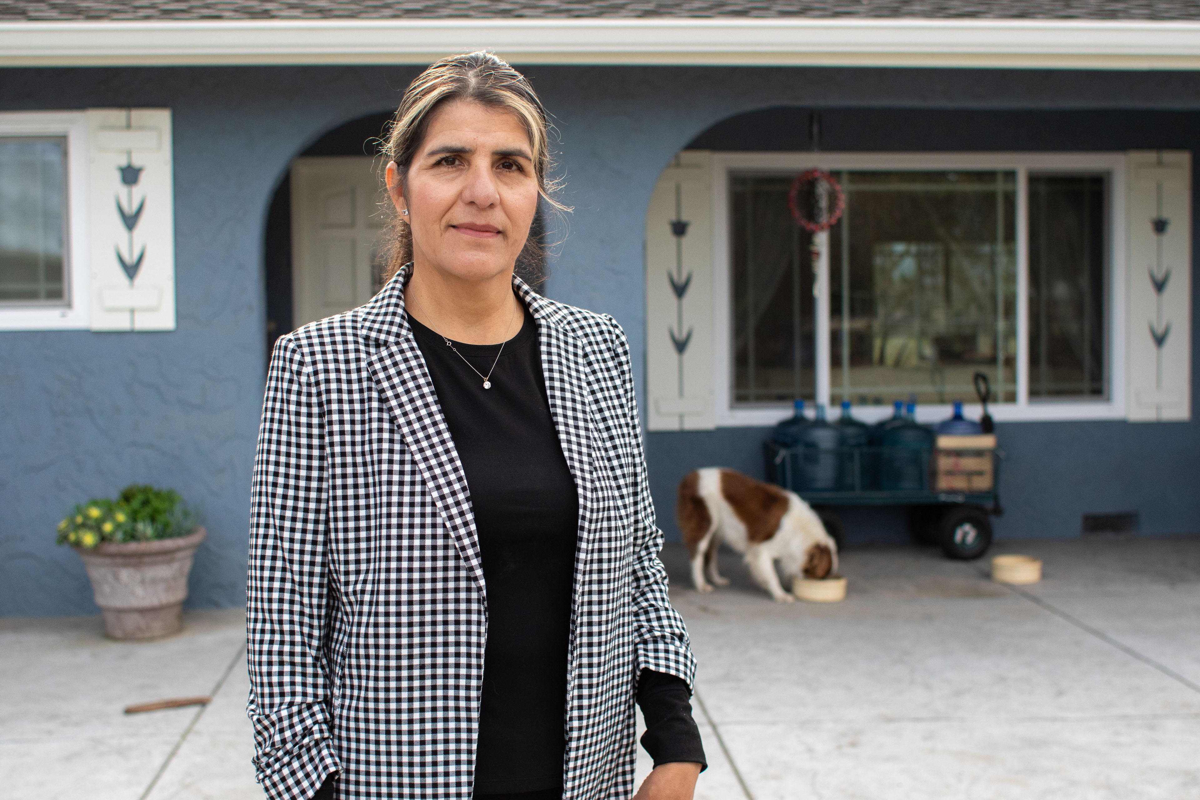 A portrait of a middle-aged woman wearing a black and white checked blazer over a black shirt. She is standing outside her home; a dog can be seen eating from a bowl in the background.