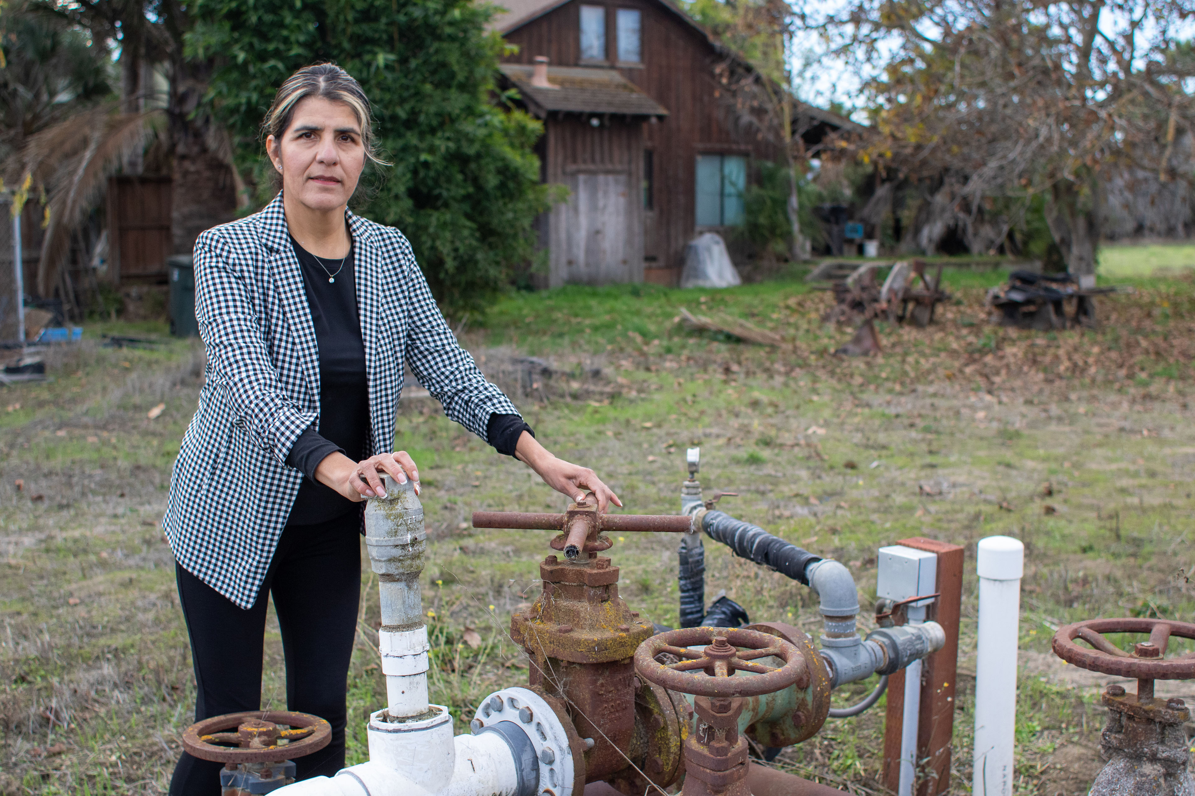 A portrait of a woman standing outside on her farm. Her hands rest on above-ground water pipes. Some are rusted and others are made of white plastic.