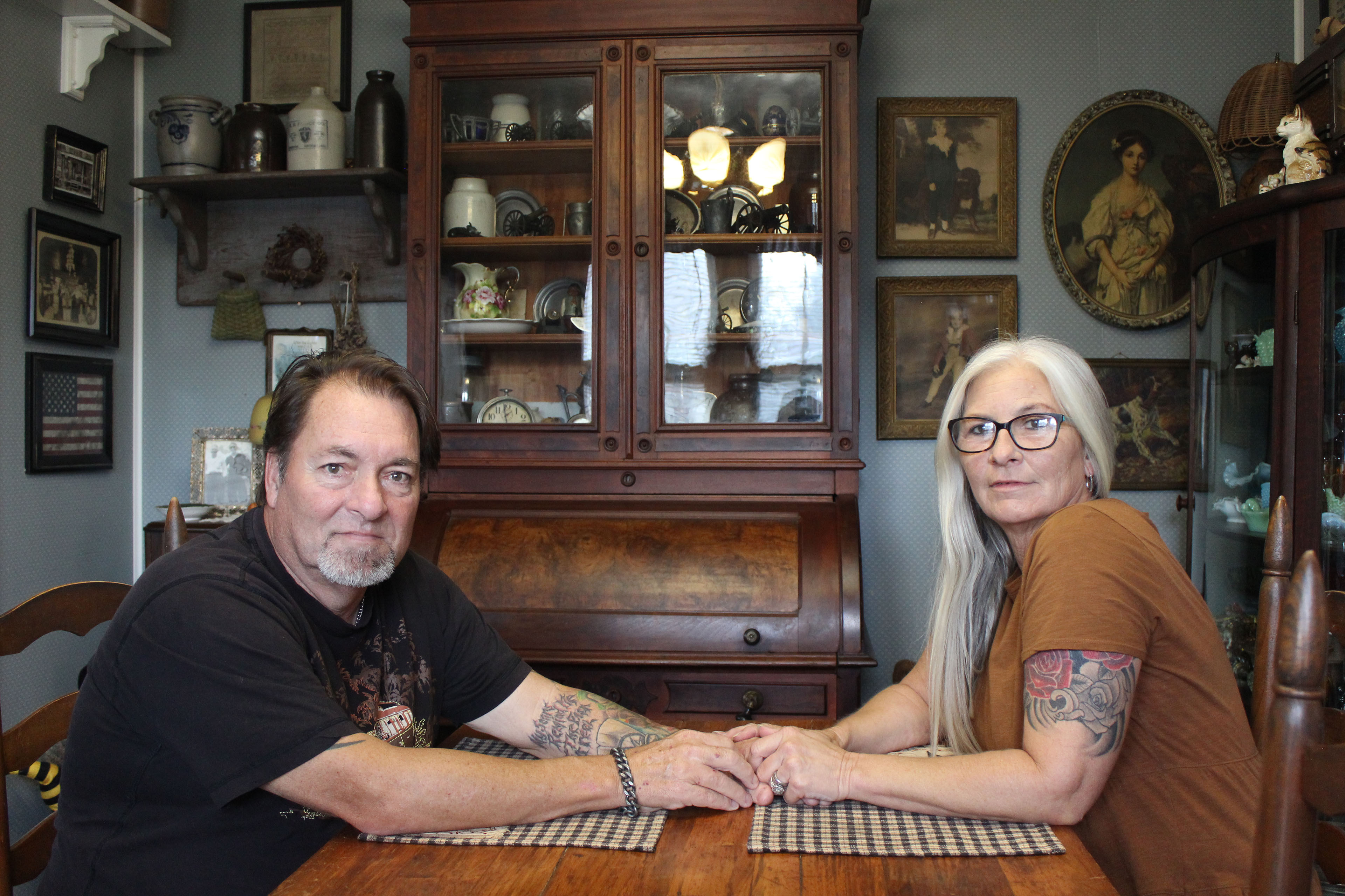 A husband and wife sit across from each other in their home dining room. They hold hands across the table and look towards the camera.