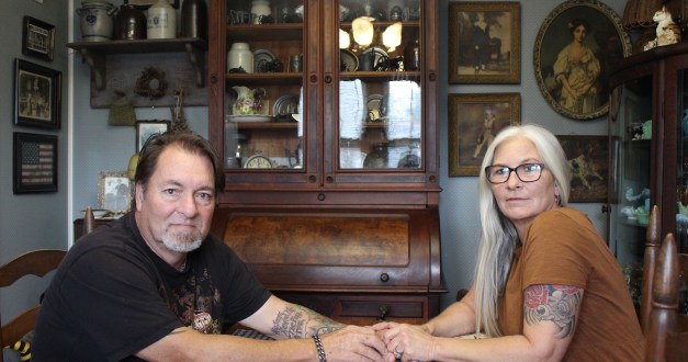 A husband and wife sit across from each other in their home dining room. They hold hands across the table and look towards the camera.