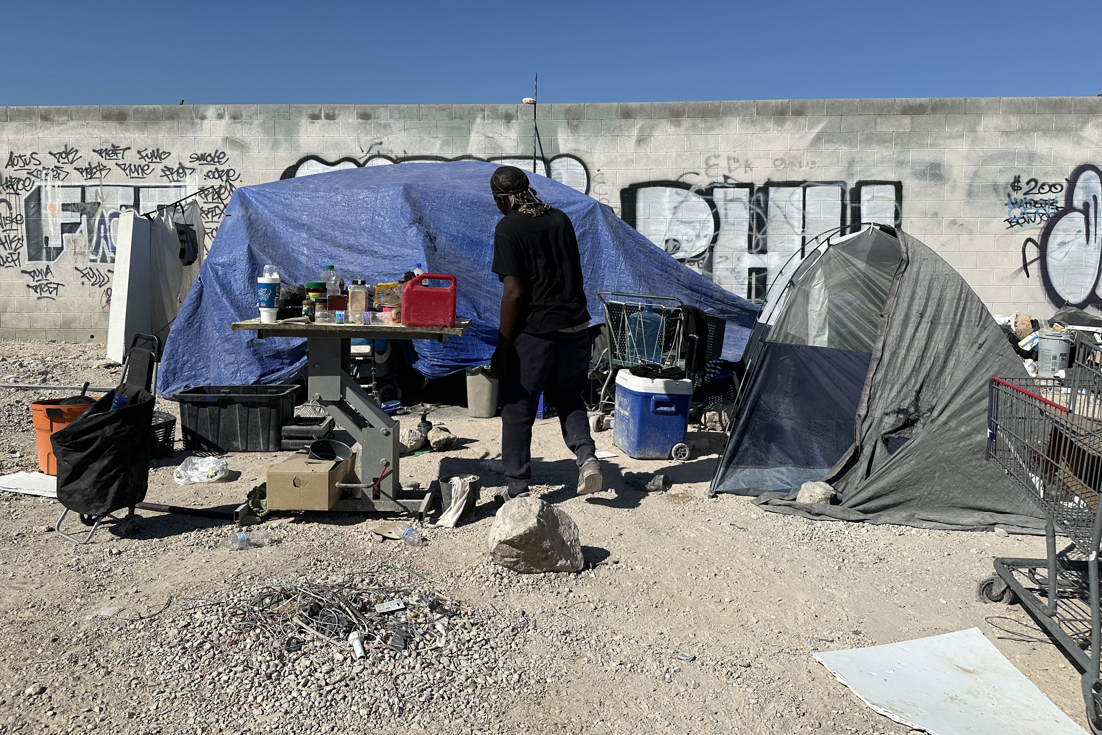 A man, with his back to the camera, walk towards his tent in a sandy area. Other personal belongings are beside his tent.