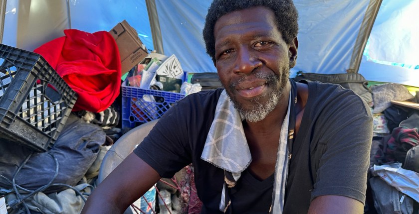 A portrait of Maurice Clark, who is sitting amongst his belongings. He has a warm expression as he looks towards the camera.