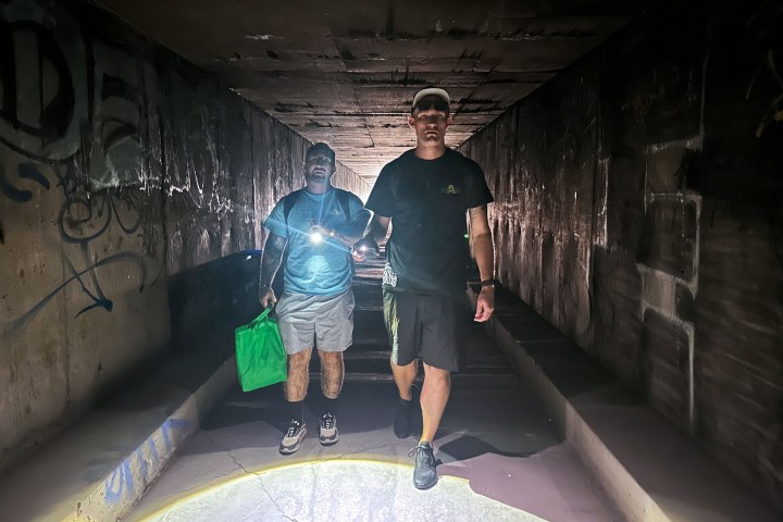 Two men walk through an underground tunnel made of concrete. They face the photographer and shine a flashlight in front of them as they walk.