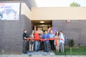 A photo showing a group of people standing in front of a brick building, behind a red ribbon, for an opening ceremony on a sunny day.