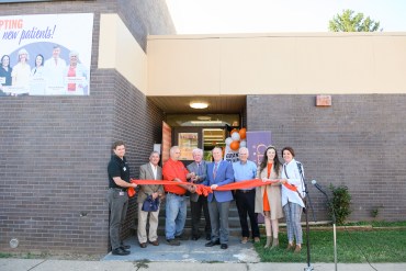 A photo showing a group of people standing in front of a brick building, behind a red ribbon, for an opening ceremony on a sunny day.