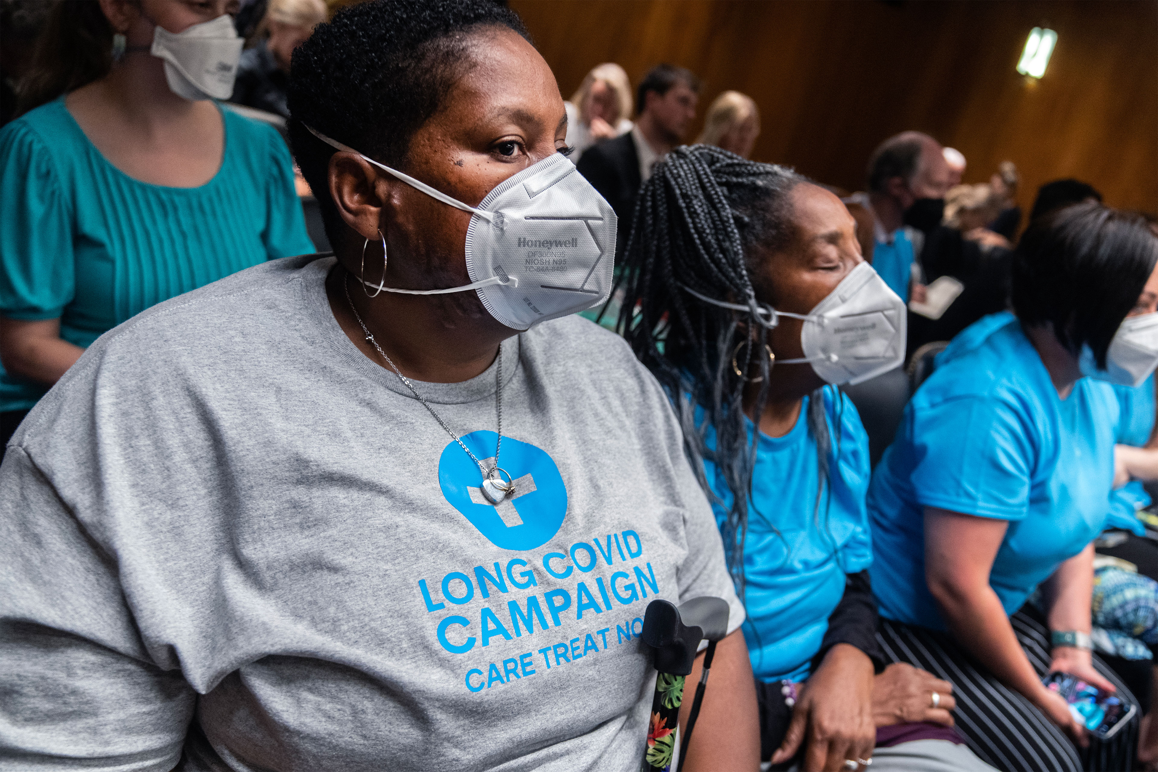 A row of women wearing N95 masks are seated. The woman in the foreground is wearing a grey t-shirt that reads "Long Covid Campaign. Care Treat Now." The women seated beside her in the same row are wearing light blue t-shirts.