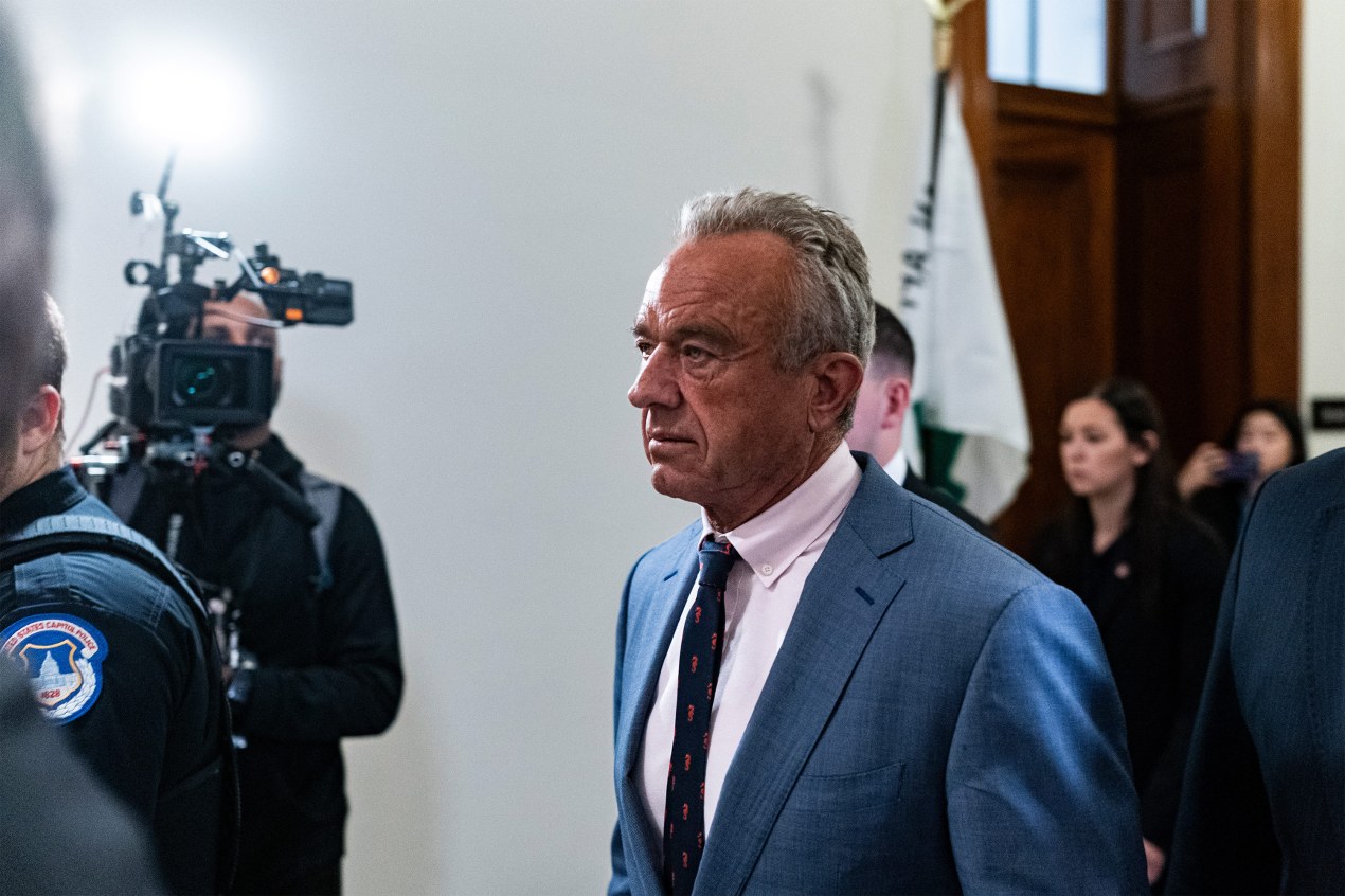 A photo of Robert F. Kennedy Jr. walking down a hallways surrounded by reporters and a TV news camera.